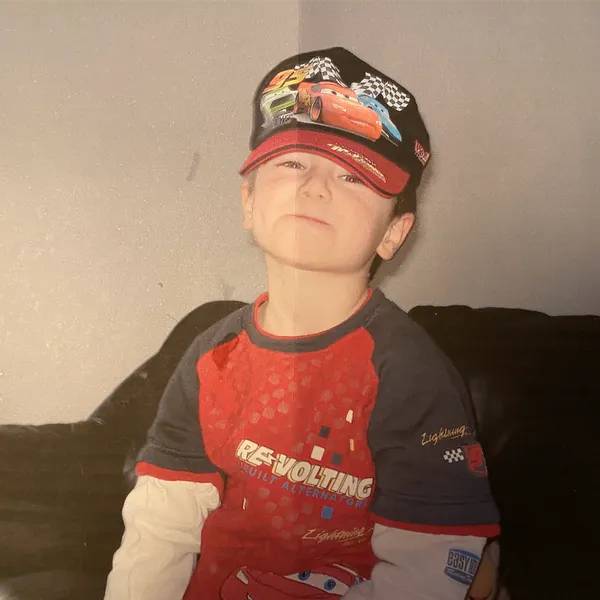 Pius as a young boy in a Cars cap and T-shirt, grinning on a sofa